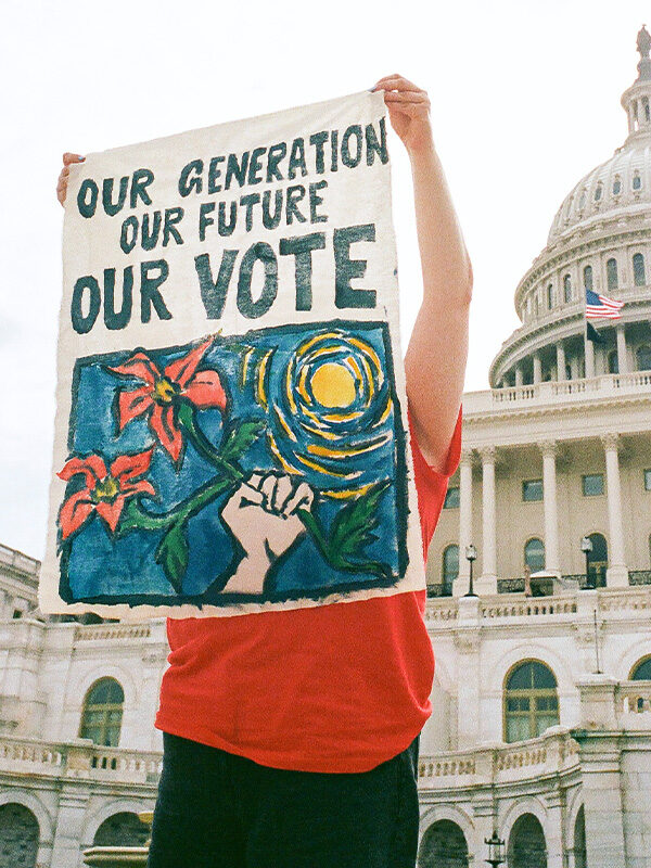 A student stands in front of the United States Capitol Building, holding a hand-painted poster that reads