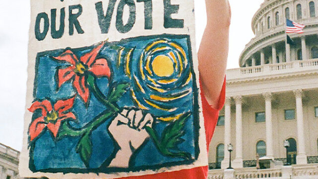 A student stands in front of the United States Capitol Building, holding a hand-painted poster that reads 