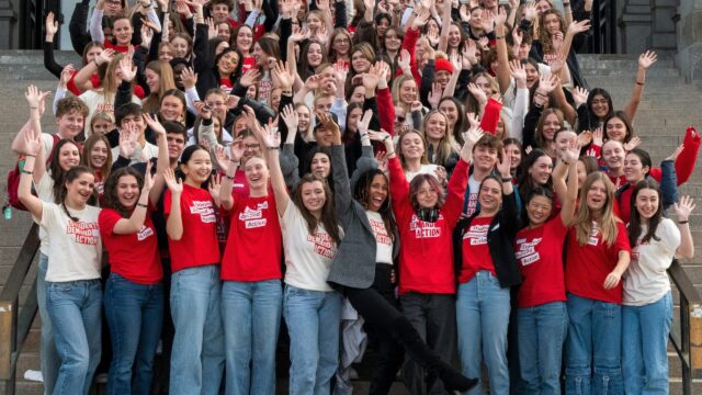 Angela Ferrell-Zabala, executive director of Moms Demand Action and Students Demand Action, stands on the steps of the Colorado State Capitol surrounded by over 100 Students Demand Action volunteers. Students are celebrating with their arms up in the air; many are wearing jeans and cream or red Students Demand Action t-shirts.