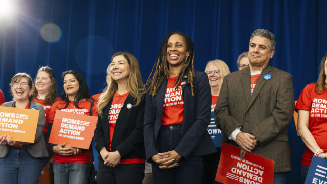 Moms Demand Action volunteers wear red t-shirts and blazers, holding Moms Demand Action signs, as they smile on stage at a rally. Angela Ferrell-Zabala, executive director of Moms Demand Action, smiles at the center of the photo.