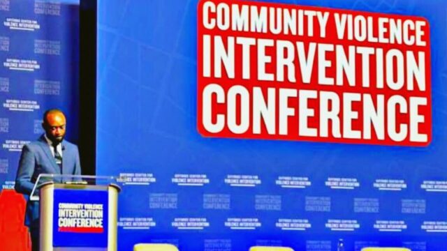Michael-Sean Spence, a Black man wearing a blue suit, light collared shirt, and a tie, presents at a podium in front of a screen that reads 